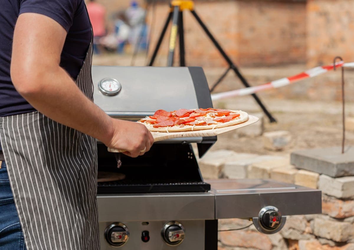 A gas-fired outdoor pizza oven on a patio with a pizza being placed inside